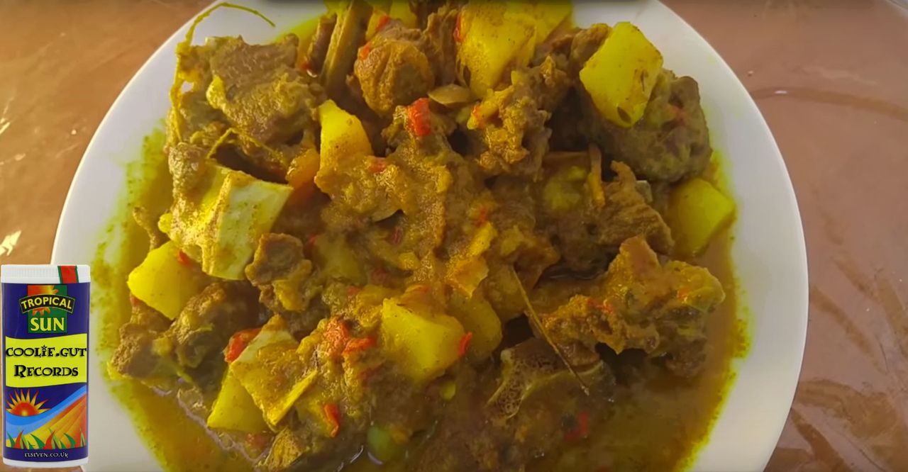 A bowl of Jamaican curry goat with a coolie.gut records juice carton beside it